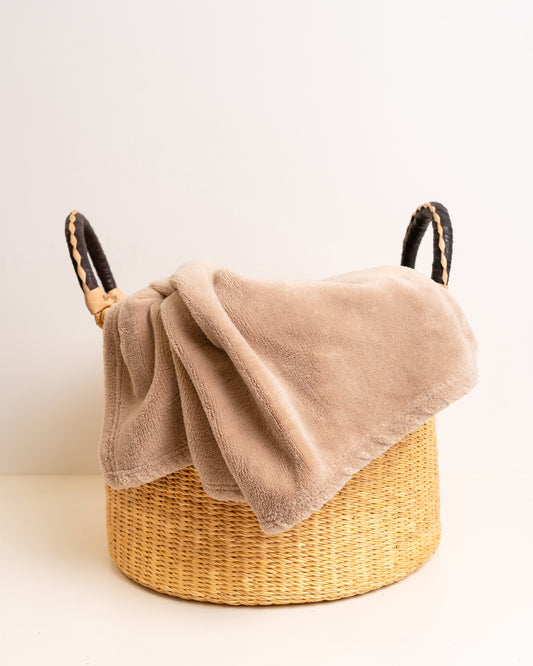 Large handwoven Bolga elephant grass basket with a draped taupe fleece blanket, styled as a living room blanket storage basket — Nathez Out of Africa