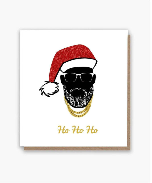 Black Santa Christmas card with sunglasses, a red Santa hat, gold chains and ‘Ho Ho Ho’ lettering, shown with a kraft envelope.