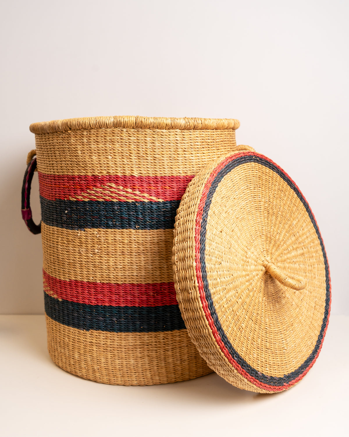 Bolga laundry basket with lid propped against it - red navy striped elephant grass storage hamper, Ghana handwoven
