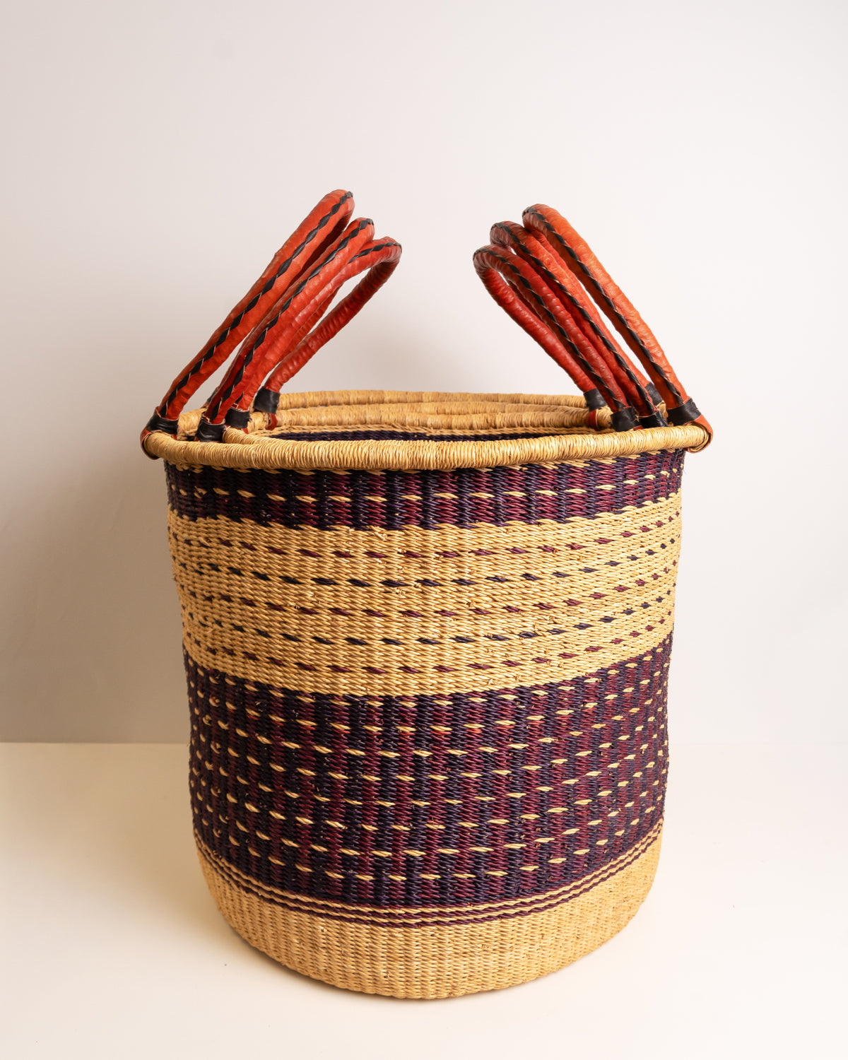 Small Bolga laundry basket in burgundy and tan stripe with leather handles standing against neutral background - handwoven elephant grass storage basket, Ghana