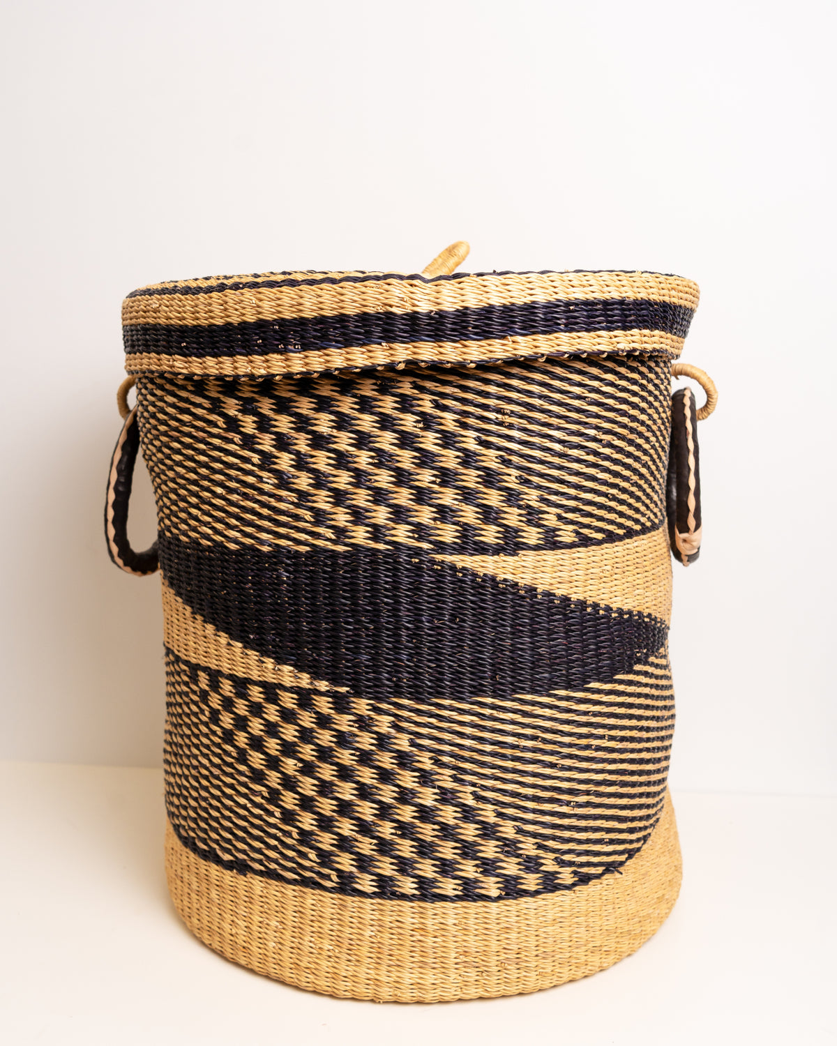 Bolga laundry basket with lid on, front angle showing black and tan woven pattern and leather handles, Ghana