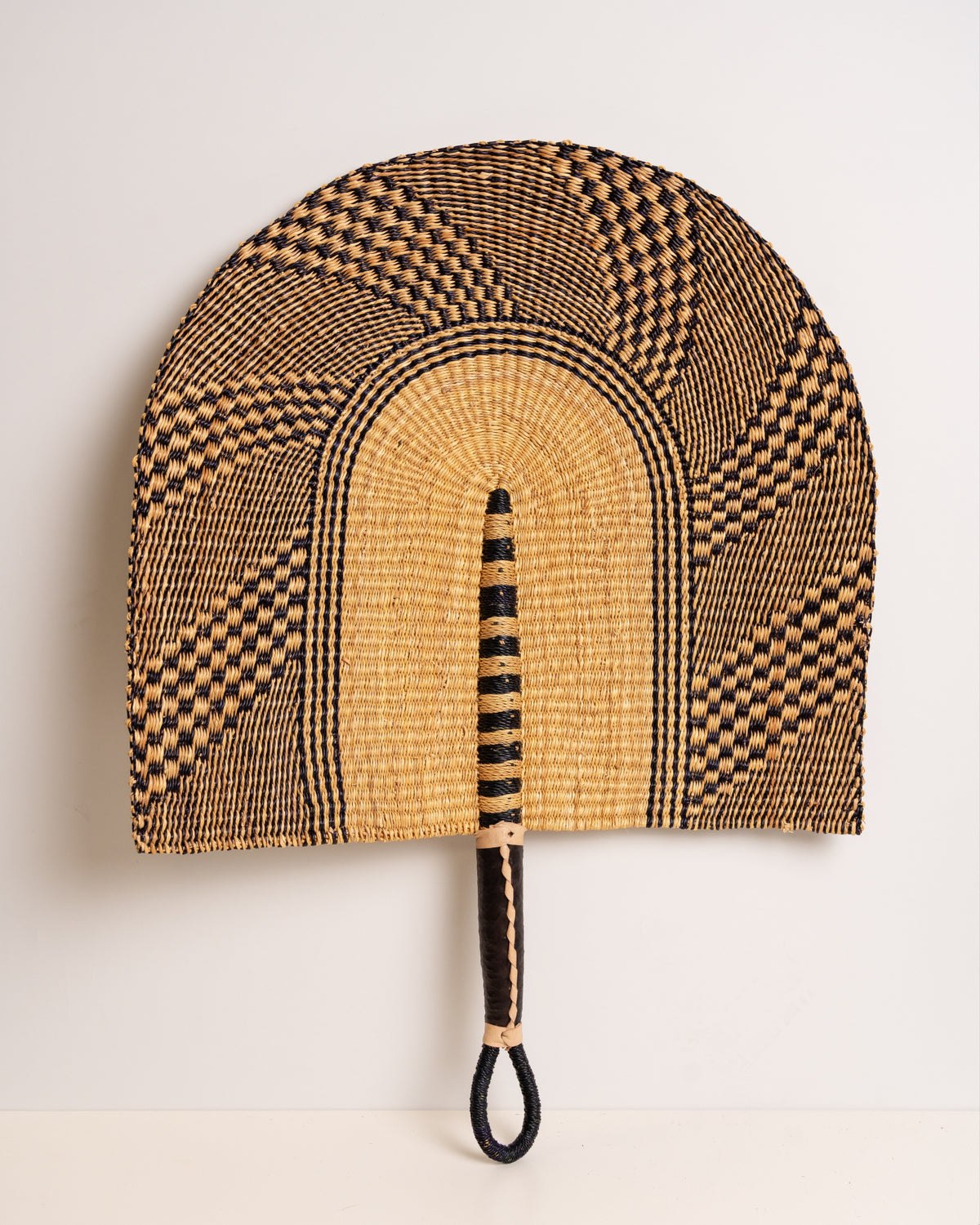 Large handwoven Bolga wall fan in natural and black elephant grass with a black leather-wrapped handle, Ghana