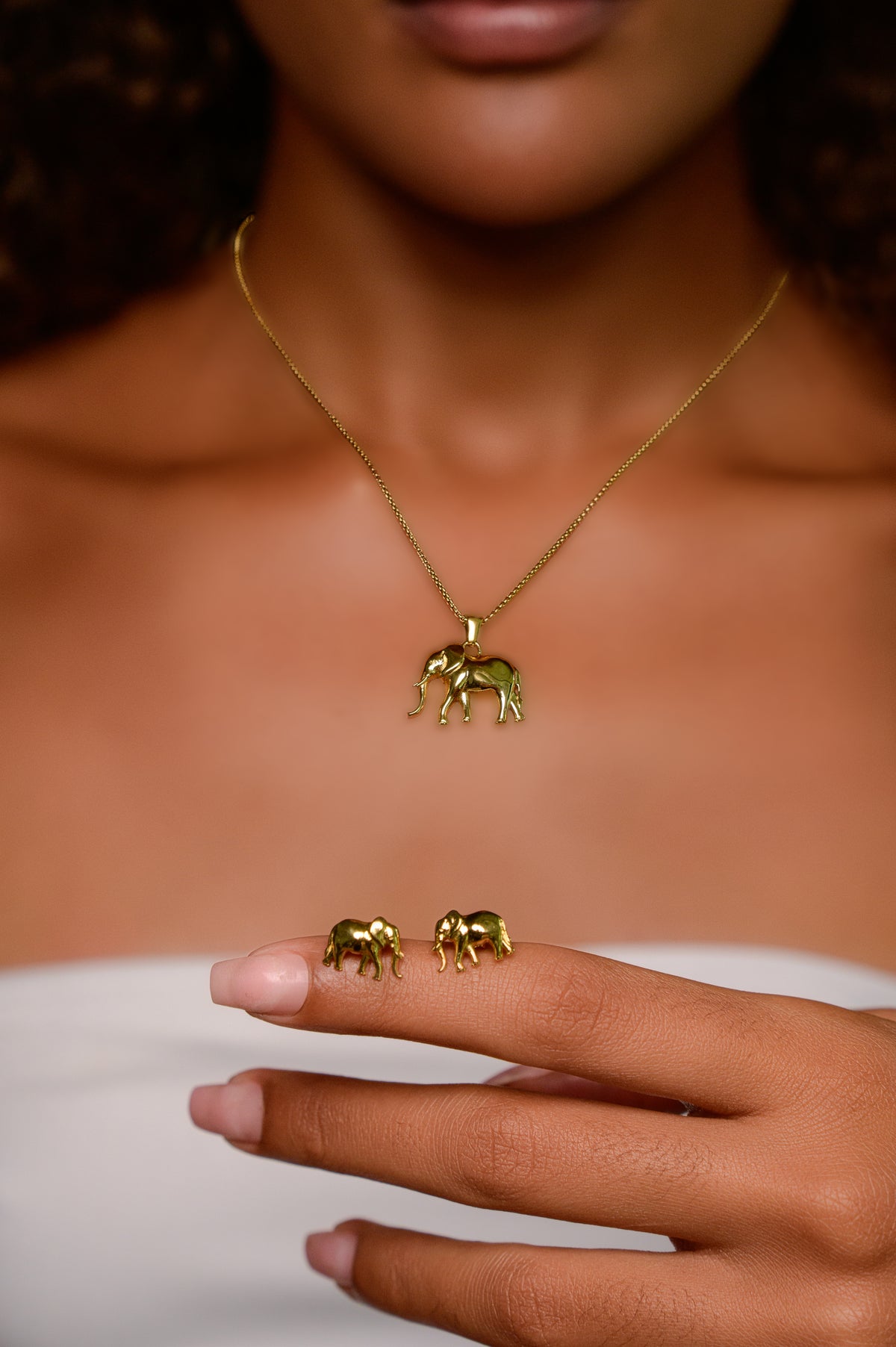 Woman wearing the Big 5 gold-plated elephant pendant necklace, holding the matching gold elephant stud earrings on her fingertip.