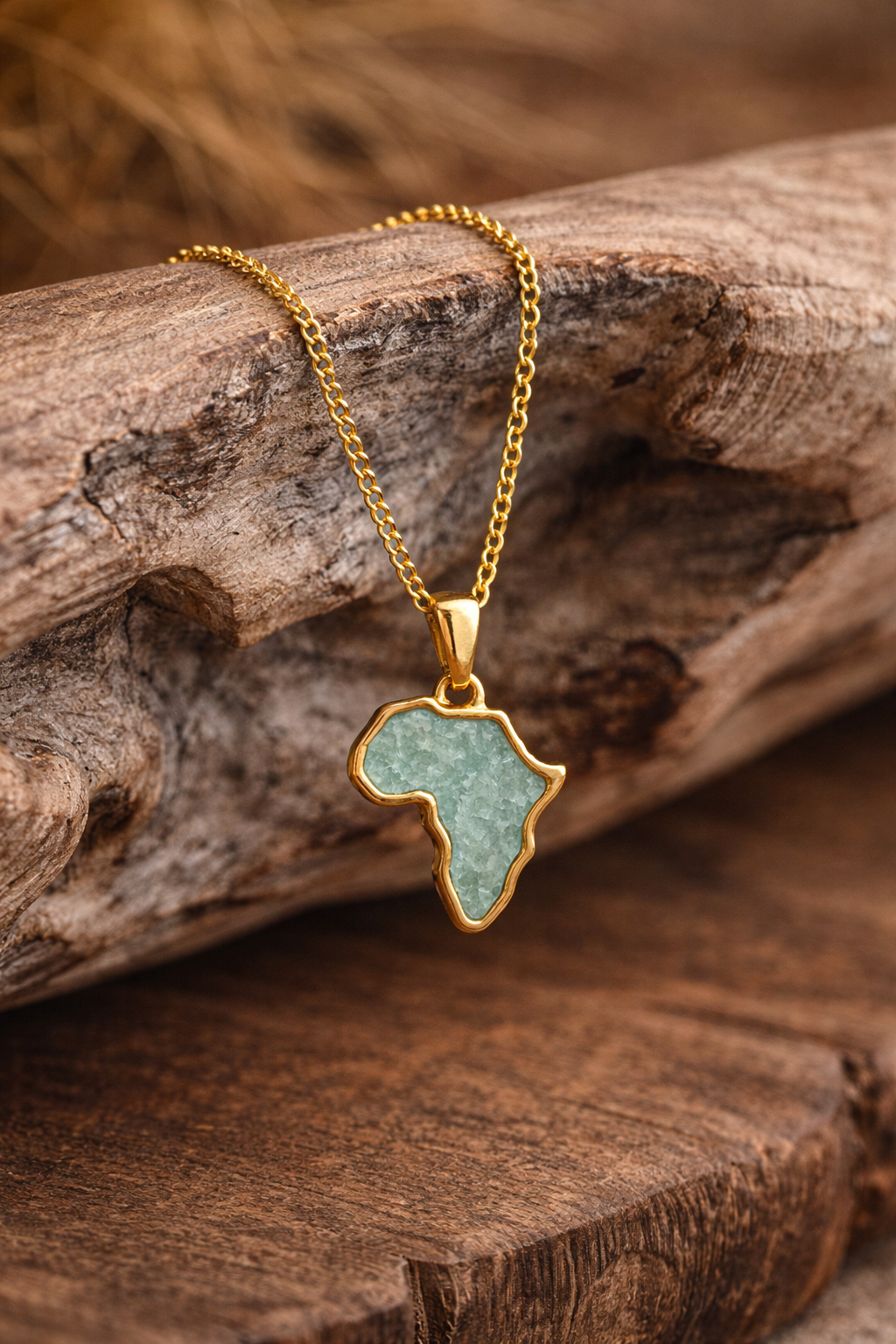 Gold-plated Africa map necklace with sea-green amazonite inlay, styled on a driftwood branch with dried grasses in the background.