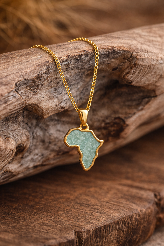 Gold-plated Africa map necklace with sea-green amazonite inlay, styled on a driftwood branch with dried grasses in the background.
