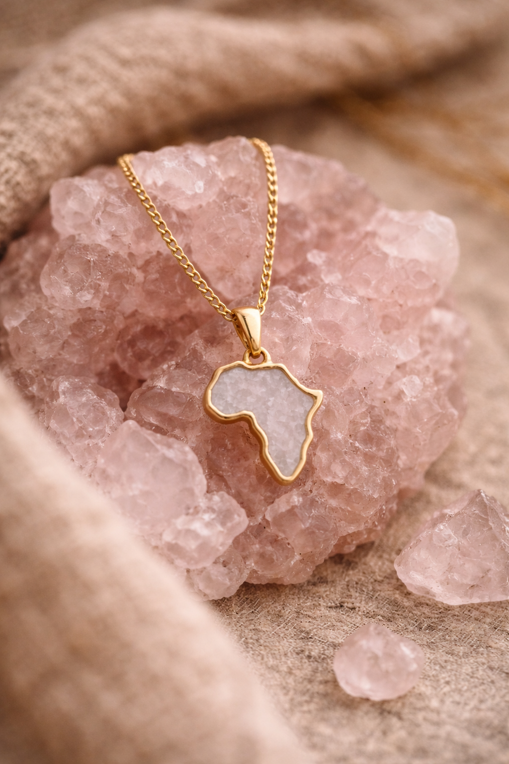 Gold-plated Africa map necklace with rose quartz inlay, styled on a raw rose quartz crystal cluster with soft beige linen.