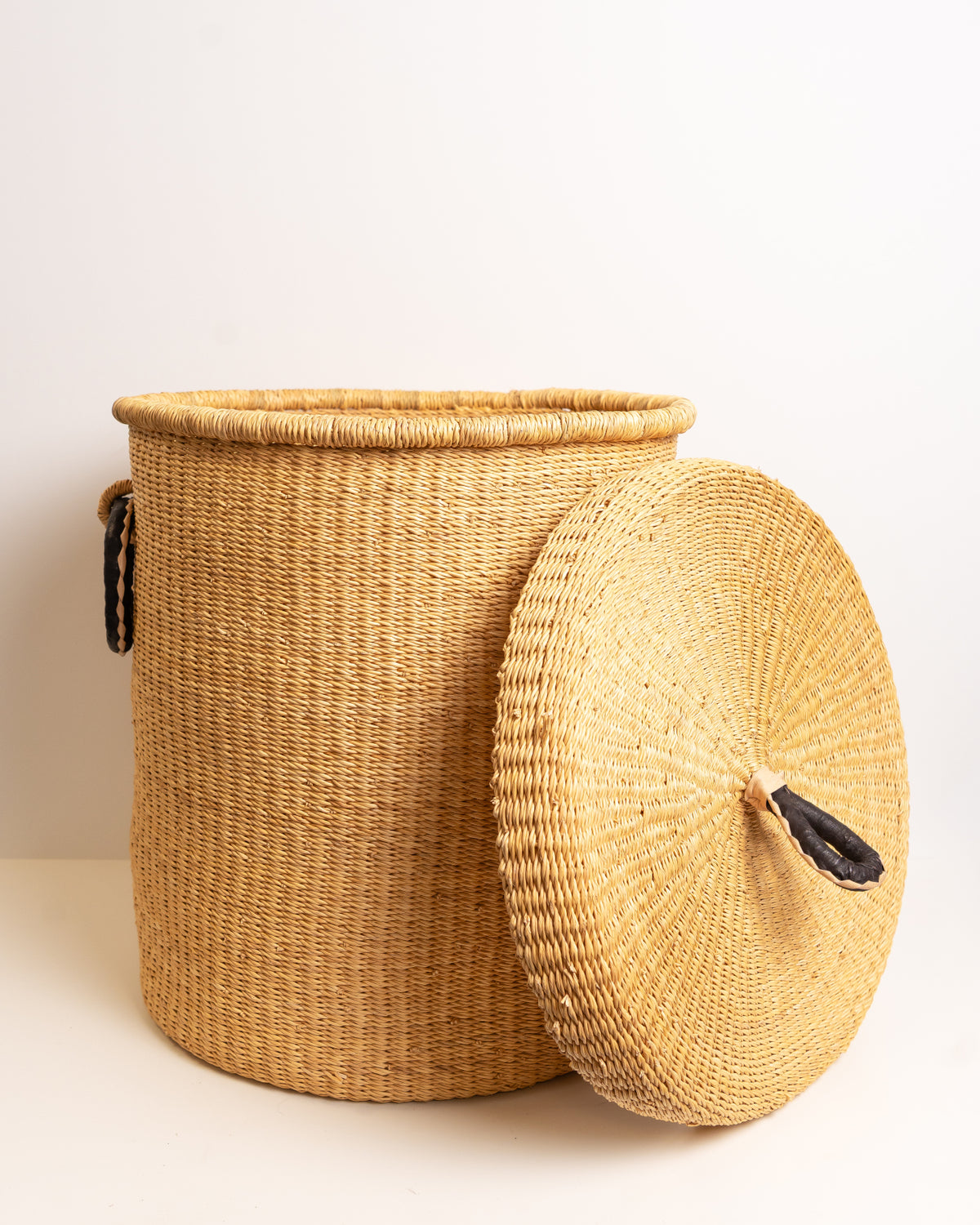 Natural tan Bolga laundry basket with lid propped against the basket, showing the woven lid underside and leather handle, handwoven elephant grass, Ghana