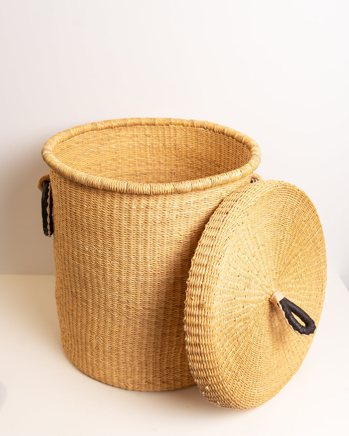 Natural tan Bolga laundry basket with lid resting beside it, showing the open rim and leather loop handles, handwoven elephant grass, Ghana
