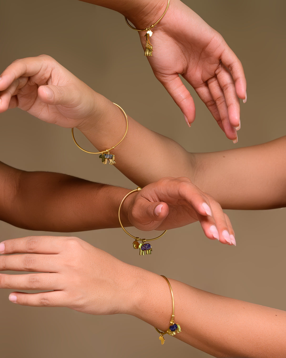 Four wrists wearing Safari Njema gold-plated charm bangles, including the giraffe bangle and elephant charm styles from the range, against a warm beige background.