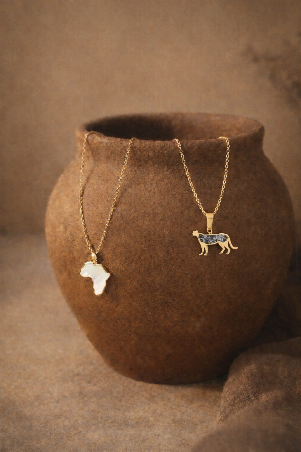 Safari Njema gold cheetah pendant necklace with snowflake obsidian inlay hanging on a clay pot beside a gold Map of Africa necklace