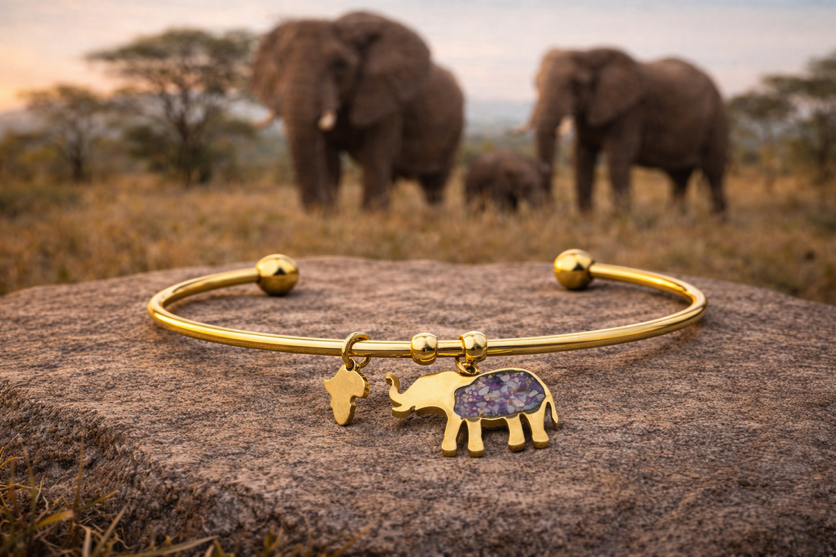 Gold-plated open cuff elephant bangle with a purple imperial jasper elephant charm and a small Africa-shaped charm, resting on a rock with elephants on the African savanna behind.