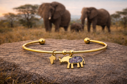 Gold-plated open cuff elephant bangle with a purple imperial jasper elephant charm and a small Africa-shaped charm, resting on a rock with elephants on the African savanna behind.