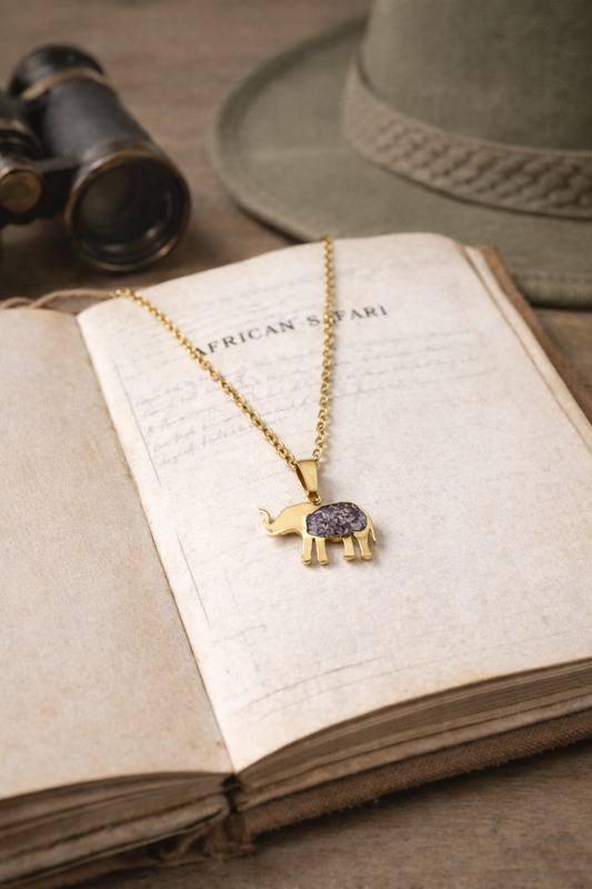 Safari Njema gold-plated elephant pendant necklace with purple imperial jasper inlay, resting on an open African Safari book beside binoculars and a safari hat.