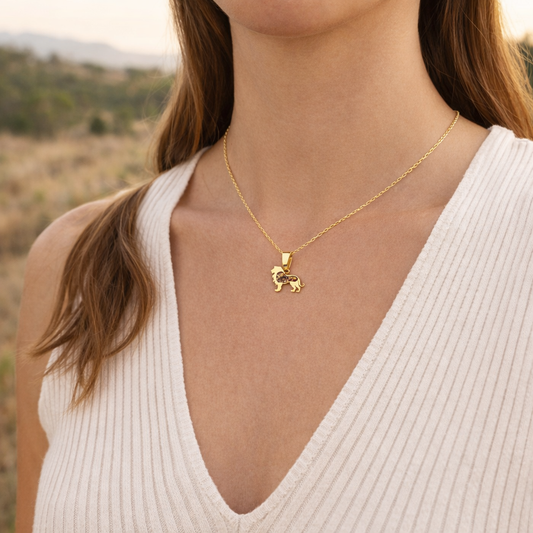 Woman in a white knit top wearing the Safari Njema gold-plated lion pendant necklace with mookaite inlay, against a soft golden savanna backdrop.